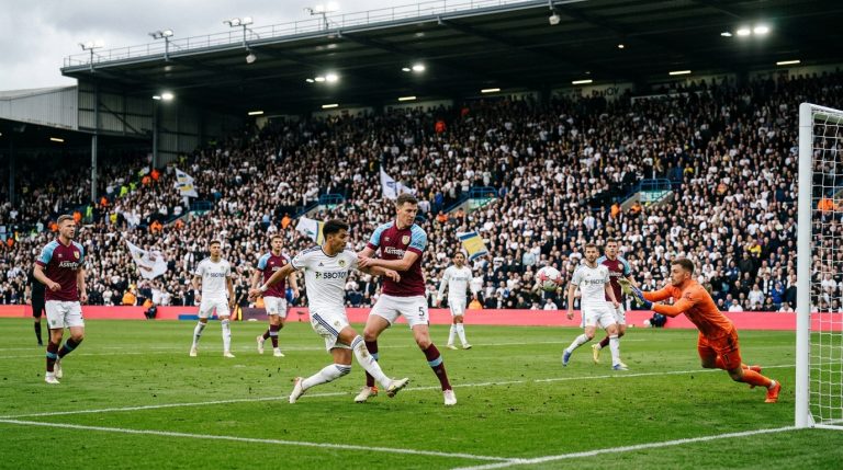 Soccer match in progress: white and maroon teams contest near the goal as the goalkeeper dives to block a shot.