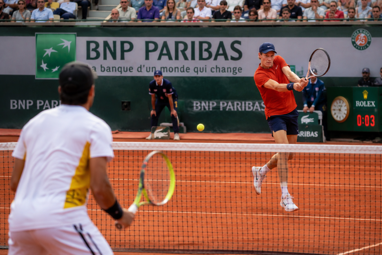 Tennis match on a red clay court with BNP Paribas banners; a player in orange shirt and cap hits a backhand toward the net.