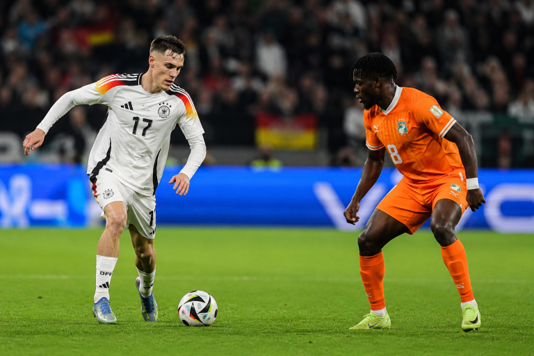 Two soccer players chase the ball on a lush green field during a match, one in a white Germany kit (number 17) and the other in an orange Netherlands kit (number 8).