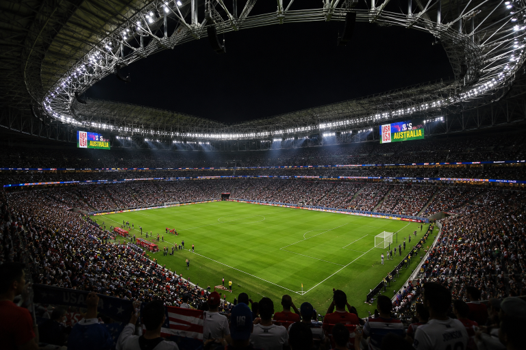 Nighttime view of a packed soccer stadium with a bright green pitch and spectators, scoreboard reading USA vs Australia.